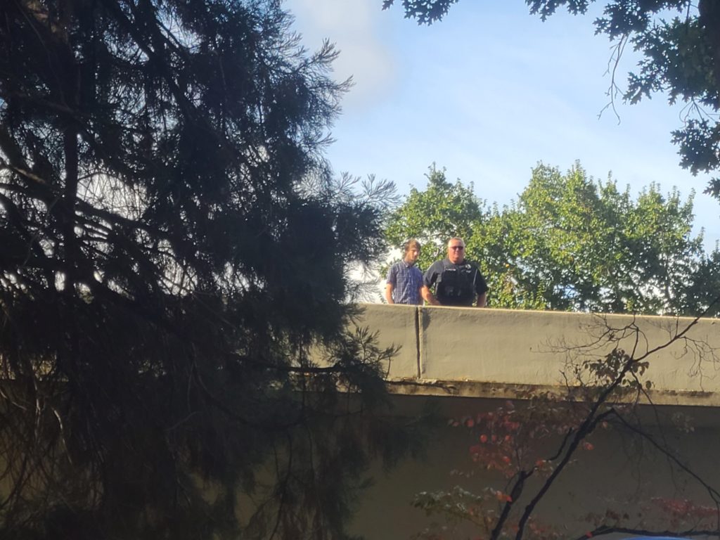 A uniformed officer stands on the overpass. A young looking person stands next to him.